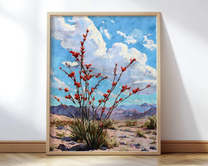 Framed desert landscape painting featuring a flowering ocotillo plant with vivid red blossoms against a bright blue sky and billowing white clouds, with distant purple mountains on the horizon. The artwork rests on a light wooden floor against a white wall, bathed in soft natural light.