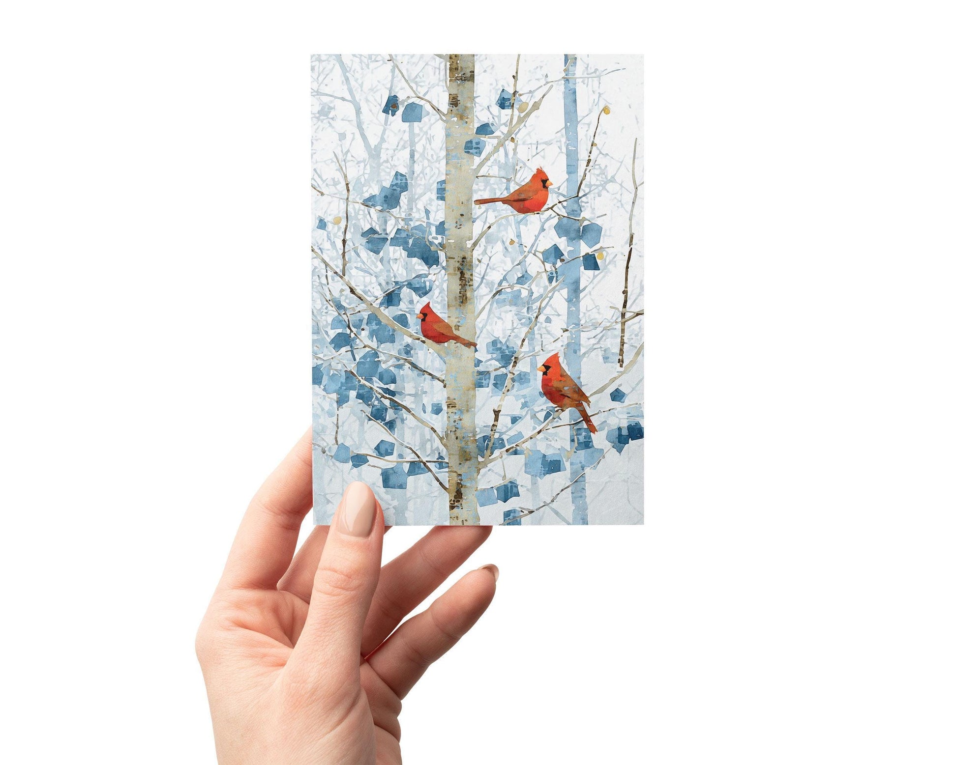 Hand holding a winter-themed greeting card featuring three bright red cardinals perched on pale branches against a soft blue and white abstract forest background. The card is shown vertically, with the artwork centered and the hand positioned at the lower left against a clean white backdrop.