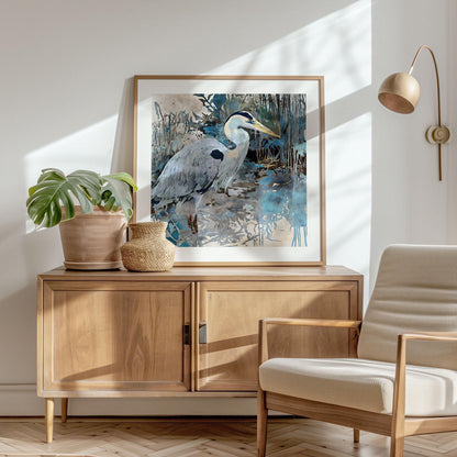Framed watercolor-style heron artwork rests on a wooden sideboard with a potted monstera and woven basket, beside a light wood and cream armchair in a sunlit modern living room with minimalist décor.