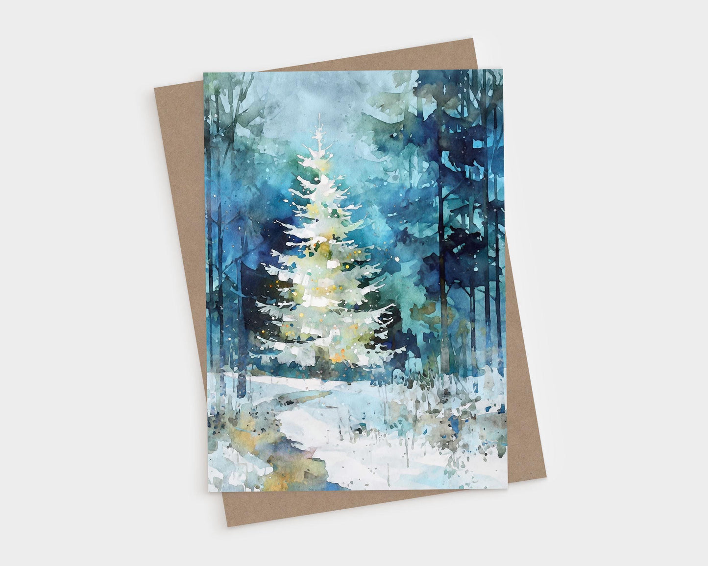 A folded greeting card featuring a watercolor-style painting of a glowing white Christmas tree in a snowy blue-green forest, shown with a kraft envelope. Soft washes, tall tree silhouettes, and gentle light speckles create a serene, wintry holiday scene.