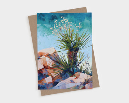 Greeting card featuring a colorful desert landscape painting of a yucca plant with spiky green leaves and small white blossoms, set against a vivid blue sky and warm-toned rocky terrain. Card is shown with a brown kraft envelope.